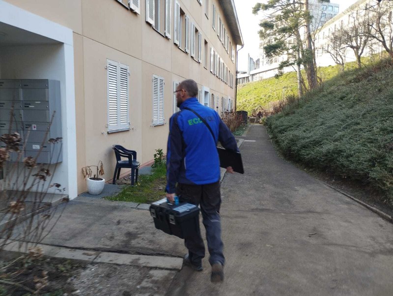 Un technicien Ecotox marche le long d&rsquo;un immeuble dans le canton du Valais, portant une mallette et du mat&eacute;riel pour une intervention de lutte antiparasitaire.